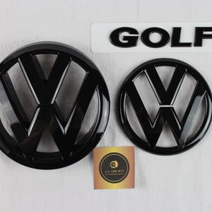 Puede incluir: Dos emblemas circulares de Volkswagen en color negro brillante de diferentes tamaños y un emblema GOLF negro. Una pequeña tarjeta cuadrada con un logotipo circular y el texto ALL CAR BITS se sitúa entre los dos emblemas sobre una superficie blanca.
