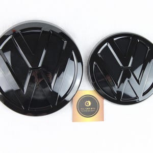 Peut inclure: Deux emblèmes Volkswagen circulaires en noir brillant de tailles différentes, présentant les lettres V et W en relief. Une petite carte carrée avec le texte ALL CAR BITZ est placée entre les deux emblèmes sur un fond blanc uni.