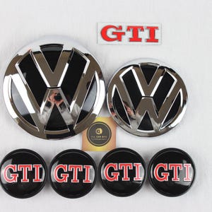 May include: Two circular Volkswagen emblems with a chrome and black finish, four smaller circular wheel center caps featuring the red and white GTI logo on a black background, and one rectangular GTI emblem with red letters on a white background.