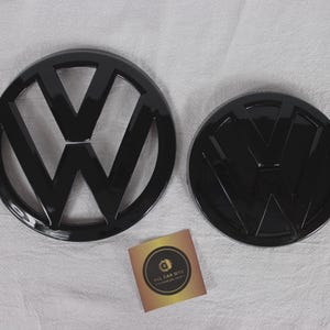 Puede incluir: Dos emblemas Volkswagen de color negro brillante, uno con marco abierto y otro con respaldo sólido, junto a una pequeña tarjeta cuadrada con un logotipo circular y el texto ALL CAR BITZ en un diseño degradado dorado y negro sobre tela blanca.