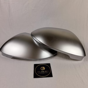 Puede incluir: Dos cubiertas de espejo lateral para automóvil de color plateado con un acabado metálico liso. Una pequeña tarjeta cuadrada negra está colocada frente a las cubiertas, con un logotipo dorado y el texto ALL CAR BITZ You know you need it.