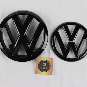 Puede incluir: Dos emblemas circulares de Volkswagen en negro brillante de diferentes tamaños, con el icónico diseño del logotipo VW. Una pequeña tarjeta cuadrada con un logotipo circular dorado y negro y el texto ALL CAR BITZ se sitúa entre ambos.