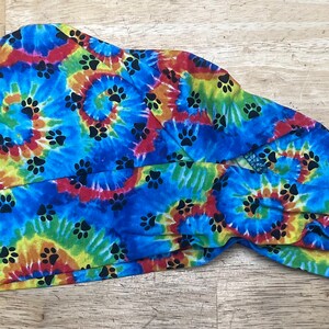 May include: A tie-dye patterned head covering in shades of blue, yellow, red, and green. The fabric features a spiral design with black paw prints. The head covering is tied at the back.