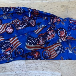 May include: A blue head wrap featuring a repeating pattern of motorcycles and American flags. The design incorporates red, white, and blue colors. The head wrap is made of a soft fabric and is designed to be tied at the back.