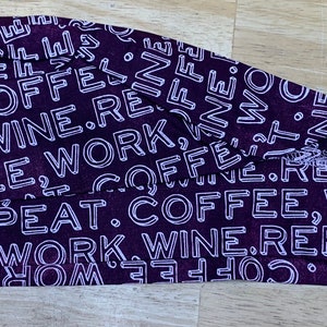May include: A dark purple surgical scrub cap with a white repeating text pattern. The text includes the words "COFFEE, WORK, WINE, REPEAT". The cap is made of a soft fabric and is designed to cover the head and hair.