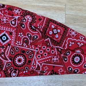 May include: A red bandana-patterned head covering with a paisley design. The fabric features a repeating pattern of white and black paisley and floral motifs on a red background. The head covering is folded and has a long, pointed tail.