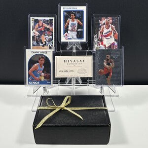 K&ouml;nnte beinhalten: Eine Sammlung von f&uuml;nf Basketball-Sammelkarten mit den Spielern Danny Ainge, Kevin McHale und anderen, pr&auml;sentiert in einem klaren, gestuften Acrylst&auml;nder neben einer Hiyasat Exhibition-Karte, hinter einer schwarzen Geschenkbox mit goldener Schleife.