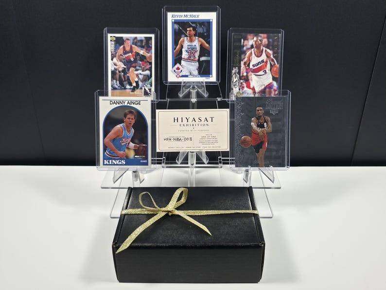 K&ouml;nnte beinhalten: Eine Sammlung von f&uuml;nf Basketball-Sammelkarten mit den Spielern Danny Ainge, Kevin McHale und anderen, pr&auml;sentiert in einem klaren, gestuften Acrylst&auml;nder neben einer Hiyasat Exhibition-Karte, hinter einer schwarzen Geschenkbox mit goldener Schleife.