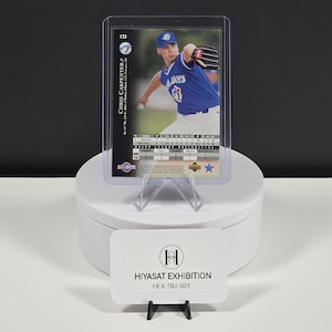 Puede incluir: Una tarjeta de b&eacute;isbol de Chris Carpenter en una funda protectora de pl&aacute;stico, con el jugador en un uniforme azul de los Toronto Blue Jays. La tarjeta muestra estad&iacute;sticas, el n&uacute;mero 133 y el logo. Un letrero blanco dice HIYASAT EXHIBITION HEX-TBJ-001.