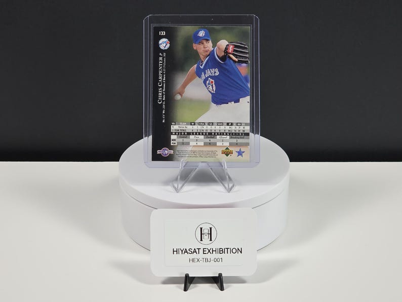Puede incluir: Una tarjeta de b&eacute;isbol de Chris Carpenter en una funda protectora de pl&aacute;stico, con el jugador en un uniforme azul de los Toronto Blue Jays. La tarjeta muestra estad&iacute;sticas, el n&uacute;mero 133 y el logo. Un letrero blanco dice HIYASAT EXHIBITION HEX-TBJ-001.