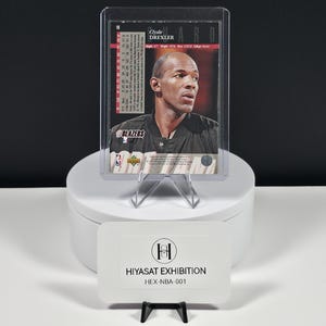 May include: A basketball trading card featuring Clyde Drexler in a black jersey, encased in a clear plastic protective sleeve. The card displays text including the player's name, team logo, and statistics. A white sign reads HIYASAT EXHIBITION HEX-NBA-001.