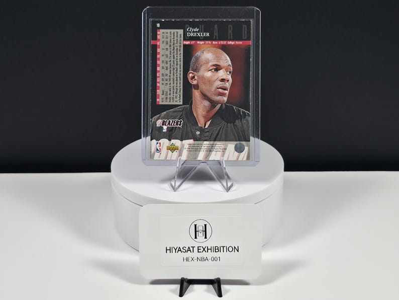 May include: A basketball trading card featuring Clyde Drexler in a black jersey, encased in a clear plastic protective sleeve. The card displays text including the player's name, team logo, and statistics. A white sign reads HIYASAT EXHIBITION HEX-NBA-001.