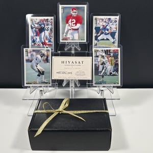 K&ouml;nnte beinhalten: Eine Sammlung von f&uuml;nf American-Football-Sammelkarten in durchsichtigen Kunststoffh&uuml;llen auf einem gestuften Acrylst&auml;nder, begleitet von einer Hiyasat Exhibition-Karte mit der Aufschrift HEX-NFL-002, hinter einer schwarzen Geschenkbox mit Goldband.