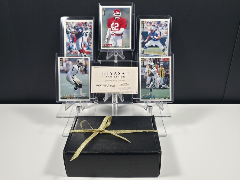 K&ouml;nnte beinhalten: Eine Sammlung von f&uuml;nf American-Football-Sammelkarten in durchsichtigen Kunststoffh&uuml;llen auf einem gestuften Acrylst&auml;nder, begleitet von einer Hiyasat Exhibition-Karte mit der Aufschrift HEX-NFL-002, hinter einer schwarzen Geschenkbox mit Goldband.