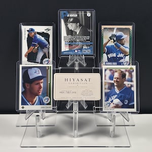 May include: A clear acrylic tiered display stand holding five baseball trading cards and a rectangular white card with the text HIYASAT EXHIBITION CURATED WITH PURPOSE HEX-TBJ-002 ONE-OF-ONE. The stand features multiple levels to showcase the cards.