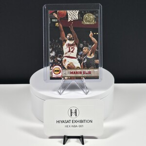 May include: A basketball trading card featuring Mario Elie in a white Rockets uniform, encased in a clear plastic protective sleeve. The card displays the NBA Hoops logo and the text Mario Elie. A white card with the text HIYASAT EXHIBITION HEX-NBA-001 sits in front.