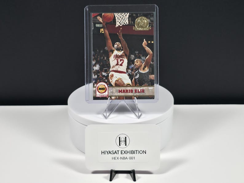 May include: A basketball trading card featuring Mario Elie in a white Rockets uniform, encased in a clear plastic protective sleeve. The card displays the NBA Hoops logo and the text Mario Elie. A white card with the text HIYASAT EXHIBITION HEX-NBA-001 sits in front.