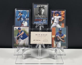 May include: A clear acrylic tiered display stand holding five baseball trading cards and a rectangular white card with the text HIYASAT EXHIBITION CURATED WITH PURPOSE HEX-TBJ-001 ONE OF ONE. The cards feature various baseball players in blue uniforms.