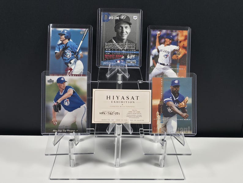 Puede incluir: Un expositor escalonado de acr&iacute;lico transparente que contiene cinco tarjetas de b&eacute;isbol y una tarjeta blanca rectangular con el texto HIYASAT EXHIBITION CURATED WITH PURPOSE HEX-TBJ-001 ONE OF ONE. Las tarjetas muestran jugadores con uniformes azules.