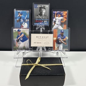 Puede incluir: Una colecci&oacute;n de cinco tarjetas de b&eacute;isbol con jugadores de los Toronto Blue Jays, exhibidas en fundas protectoras transparentes sobre un soporte de acr&iacute;lico. Una peque&ntilde;a tarjeta blanca con la etiqueta HIYASAT EXHIBITION se encuentra en el centro. Debajo hay una caja negra con una cinta dorada.