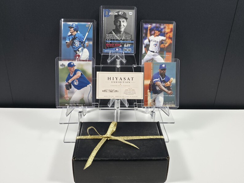 Puede incluir: Una colecci&oacute;n de cinco tarjetas de b&eacute;isbol con jugadores de los Toronto Blue Jays, exhibidas en fundas protectoras transparentes sobre un soporte de acr&iacute;lico. Una peque&ntilde;a tarjeta blanca con la etiqueta HIYASAT EXHIBITION se encuentra en el centro. Debajo hay una caja negra con una cinta dorada.