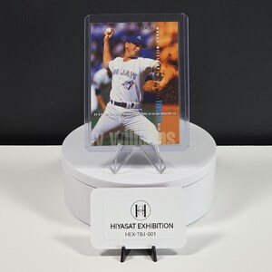 Puede incluir: Una tarjeta de b&eacute;isbol que muestra a un jugador con uniforme blanco de los Blue Jays lanzando una pelota, dentro de una funda protectora de pl&aacute;stico transparente. La tarjeta incluye el texto Woody Williams y Toronto Blue Jays. Un letrero blanco dice HIYASAT EXHIBITION.