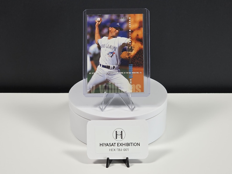 Puede incluir: Una tarjeta de b&eacute;isbol que muestra a un jugador con uniforme blanco de los Blue Jays lanzando una pelota, dentro de una funda protectora de pl&aacute;stico transparente. La tarjeta incluye el texto Woody Williams y Toronto Blue Jays. Un letrero blanco dice HIYASAT EXHIBITION.