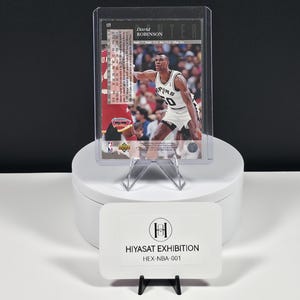 May include: A David Robinson basketball trading card in a clear protective sleeve, featuring the player in a white Spurs uniform. Below, a white rectangular sign displays the text HIYASAT EXHIBITION and the code HEX-NBA-001 in black ink.