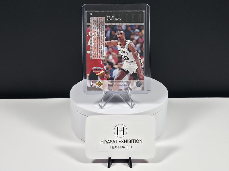 May include: A David Robinson basketball trading card in a clear protective sleeve, featuring the player in a white Spurs uniform. Below, a white rectangular sign displays the text HIYASAT EXHIBITION and the code HEX-NBA-001 in black ink.