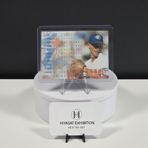Puede incluir: Una tarjeta de b&eacute;isbol en una funda de pl&aacute;stico transparente, que muestra a un jugador con gorra azul, camiseta blanca y guante marr&oacute;n. La tarjeta presenta datos estad&iacute;sticos y el texto HIYASAT EXHIBITION HEX-TBJ-001 en una etiqueta blanca.