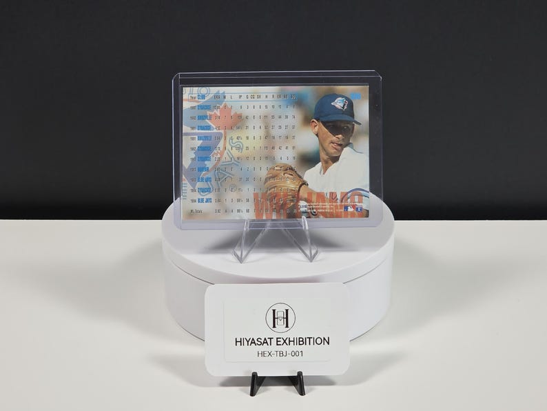 Puede incluir: Una tarjeta de b&eacute;isbol en una funda de pl&aacute;stico transparente, que muestra a un jugador con gorra azul, camiseta blanca y guante marr&oacute;n. La tarjeta presenta datos estad&iacute;sticos y el texto HIYASAT EXHIBITION HEX-TBJ-001 en una etiqueta blanca.