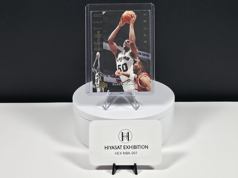 May include: A David Robinson basketball trading card in a protective plastic case, featuring the player in a white Spurs jersey with number 50, shooting a basketball. The card includes the text David Robinson and Electric Court.