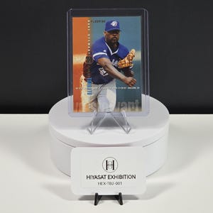 Puede incluir: Una tarjeta de b&eacute;isbol Fleer 1990 de Dave Stewart en una funda protectora transparente. La tarjeta muestra a Stewart con un uniforme azul de los Toronto Blue Jays y un guante beige. Debajo, un letrero rectangular blanco muestra el texto HIYASAT EXHIBITION HEX-TBJ-001.