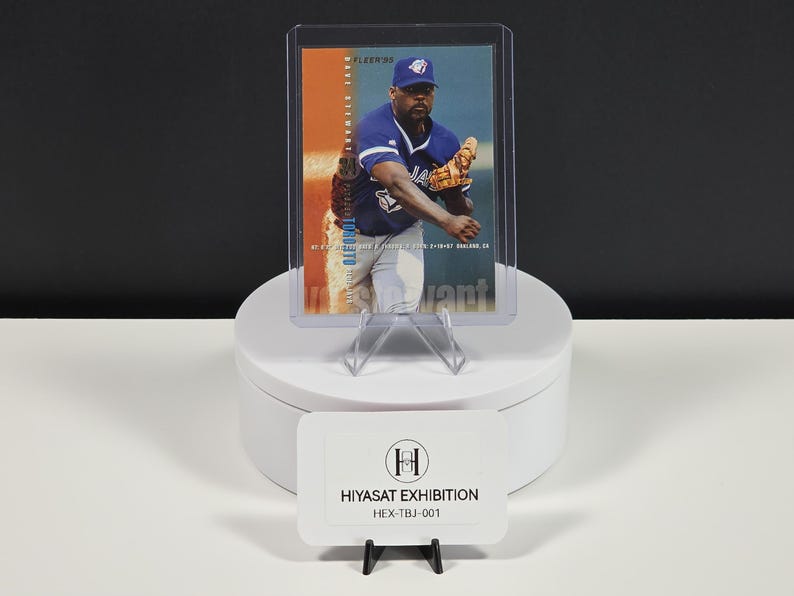 Puede incluir: Una tarjeta de b&eacute;isbol Fleer 1990 de Dave Stewart en una funda protectora transparente. La tarjeta muestra a Stewart con un uniforme azul de los Toronto Blue Jays y un guante beige. Debajo, un letrero rectangular blanco muestra el texto HIYASAT EXHIBITION HEX-TBJ-001.