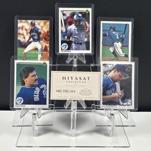 May include: A clear acrylic tiered display stand holding five baseball trading cards and a central rectangular card labeled HIYASAT EXHIBITION, HEX-TBJ-003, and ONE-OF-ONE. The cards feature Toronto Blue Jays players in blue and white uniforms.