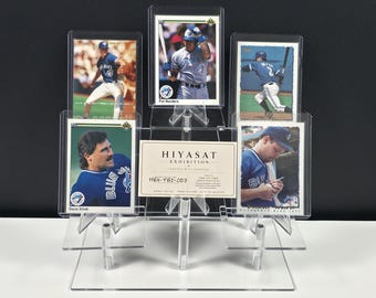 May include: A clear acrylic tiered display stand holding five baseball trading cards and a central rectangular card labeled HIYASAT EXHIBITION, HEX-TBJ-003, and ONE-OF-ONE. The cards feature Toronto Blue Jays players in blue and white uniforms.