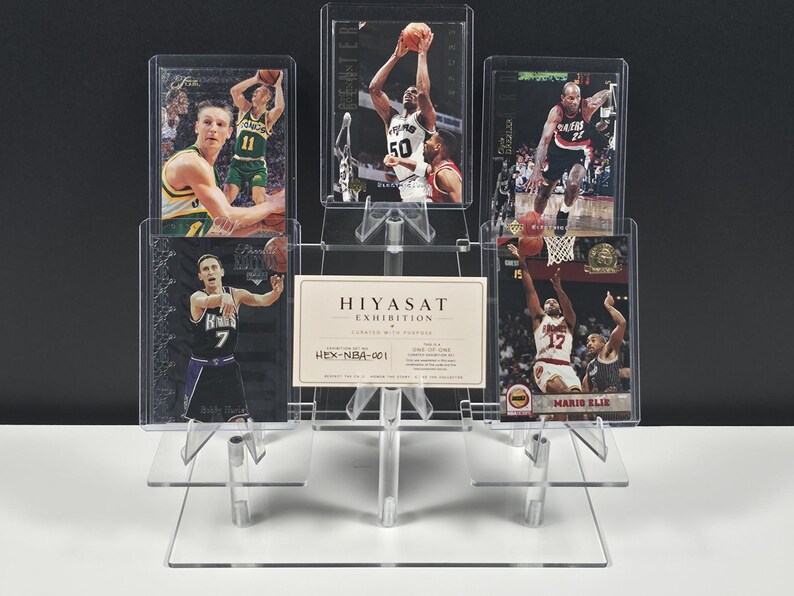 May include: A clear acrylic tiered display stand holding five basketball trading cards and a central rectangular card labeled HIYASAT EXHIBITION CURATED WITH PURPOSE HEX-NBA-001. The cards feature various players in action poses.