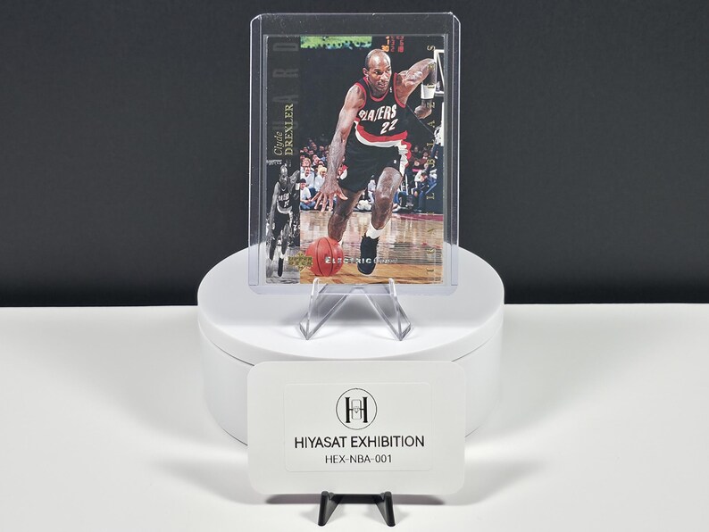 May include: A sports trading card featuring Clyde Drexler in a black and red Portland Trail Blazers uniform, dribbling a basketball on a court. The card is encased in a clear plastic protective sleeve and displays the text Clyde Drexler.