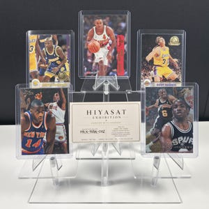 May include: A clear acrylic tiered display stand holding five basketball trading cards in protective cases. The cards feature various players in team uniforms. A rectangular white card with the text HIYASAT EXHIBITION HEX-NBA-002 is centered.