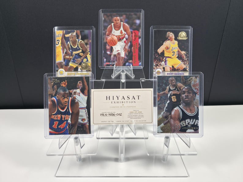 May include: A clear acrylic tiered display stand holding five basketball trading cards in protective cases. The cards feature various players in team uniforms. A rectangular white card with the text HIYASAT EXHIBITION HEX-NBA-002 is centred.