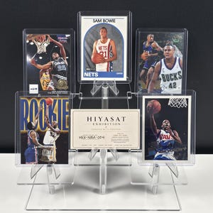 May include: A clear acrylic tiered display stand holding five basketball trading cards and a central Hiyasat Exhibition card. The cards feature various players in action, including Sam Bowie, and are encased in protective plastic sleeves.