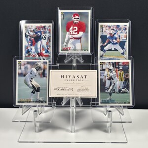 K&ouml;nnte beinhalten: Ein transparenter, abgestufter Acryl-Pr&auml;sentationsst&auml;nder mit f&uuml;nf rechteckigen Sport-Sammelkarten und einer zentralen wei&szlig;en Karte mit dem Text HIYASAT EXHIBITION CURATED WITH PURPOSE HEX-NFL-002. Die Karten zeigen verschiedene Footballspieler.