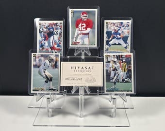 May include: A clear acrylic tiered display stand holding five rectangular sports trading cards and a central rectangular white card with the text HIYASAT EXHIBITION CURATED WITH PURPOSE HEX-NFL-002. The cards feature various football players.