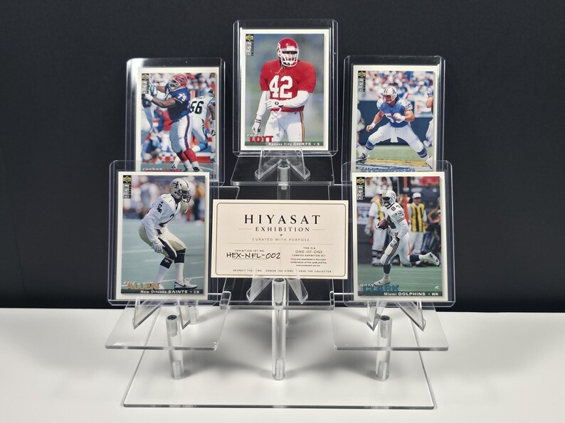 K&ouml;nnte beinhalten: Ein transparenter, abgestufter Acryl-Pr&auml;sentationsst&auml;nder mit f&uuml;nf rechteckigen Sport-Sammelkarten und einer zentralen wei&szlig;en Karte mit dem Text HIYASAT EXHIBITION CURATED WITH PURPOSE HEX-NFL-002. Die Karten zeigen verschiedene Footballspieler.