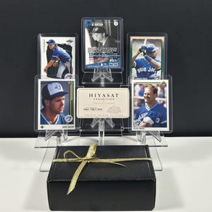 May include: A collection of five baseball trading cards displayed in a clear acrylic stand behind a black gift box tied with a gold ribbon. The cards feature players in blue uniforms, and a central white card reads HIYASAT EXHIBITION HEX-TILT-002.