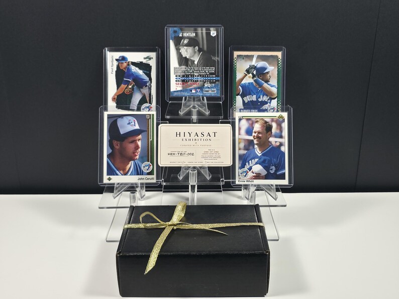 May include: A collection of five baseball trading cards displayed in a clear acrylic stand behind a black gift box tied with a gold ribbon. The cards feature players in blue uniforms, and a central white card reads HIYASAT EXHIBITION HEX-TILT-002.