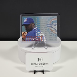 Puede incluir: Una tarjeta de b&eacute;isbol rectangular en un estuche de pl&aacute;stico transparente, con un jugador con gorra y camiseta azules. La tarjeta muestra datos estad&iacute;sticos y el logo de los Toronto Blue Jays. Un letrero blanco dice HIYASAT EXHIBITION HEX-TBJ-001.