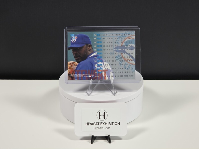 Puede incluir: Una tarjeta de b&eacute;isbol rectangular en un estuche de pl&aacute;stico transparente, con un jugador con gorra y camiseta azules. La tarjeta muestra datos estad&iacute;sticos y el logo de los Toronto Blue Jays. Un letrero blanco dice HIYASAT EXHIBITION HEX-TBJ-001.