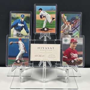 May include: A clear acrylic tiered display stand holding four baseball trading cards and a central rectangular card labeled HIYASAT EXHIBITION CURATED WITH PURPOSE. The cards feature various players in action poses, including Jim Abbott and Jim Eisenreich.