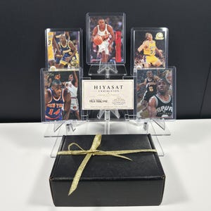 May include: A black rectangular gift box tied with a gold ribbon sits in front of a clear acrylic display stand holding five basketball trading cards and a white rectangular card with the text HIYASAT EXHIBITION and HEX-NBA-002 printed on it.